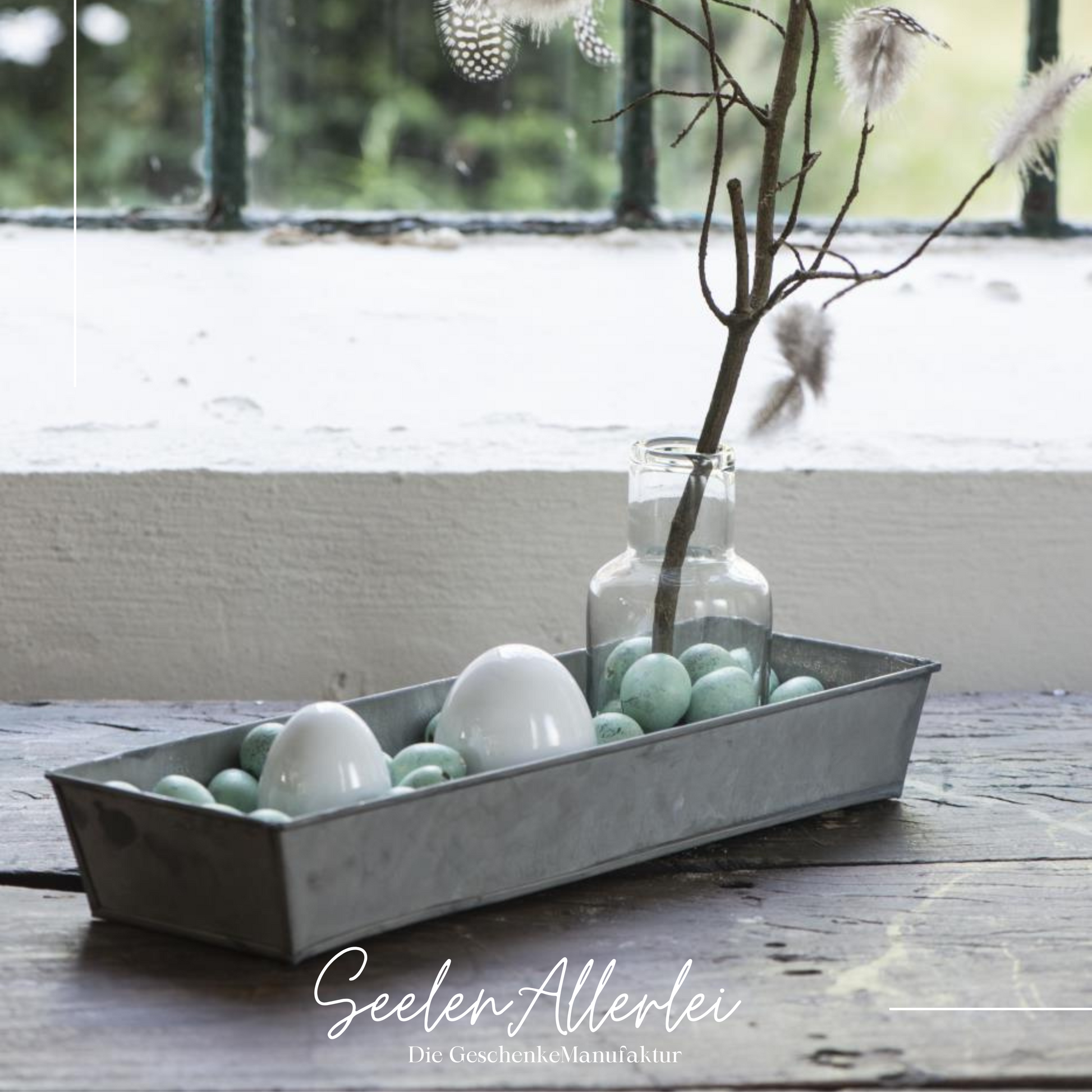 Eine Metallschale bzw ein Metalltablett steht mit Porzellan und Wachteleiern gefüllt auf einer Holzfensterbank. Neben den Eiern ist auf dem Tablett noch eine Glasvase mit einem Zweig, der mit Federn behangen ist.