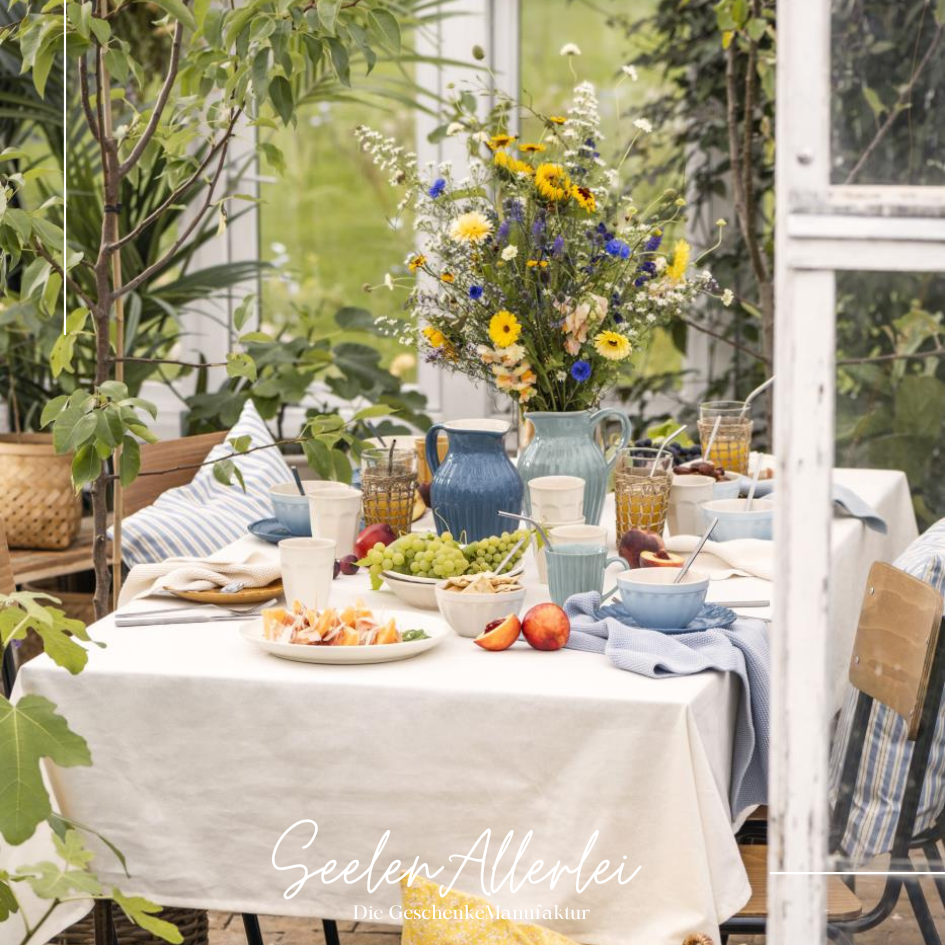 perfekt gedeckter Tisch mitweißer Tischdecke, mynte Krug von Iblaursen mit wunderschönem großen Blumenstrauß aus Sommerblumen, frischem Obst in einem Wintergarten mit vielen Grünpflanzen